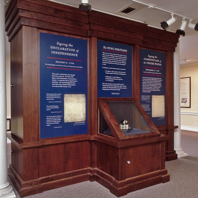 Color photo of exhibit case with text panels and silver inkstand.