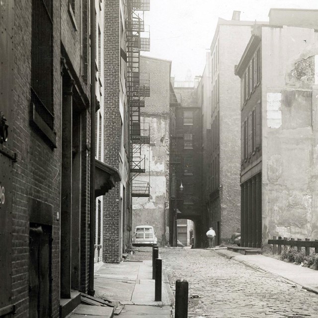Black and white photo of a narrow alley lined with multi-story buildings.