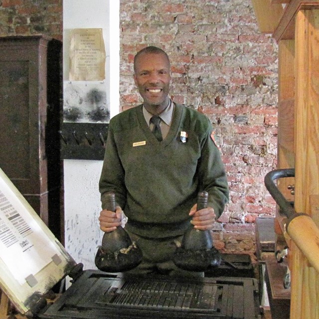 Color photo of male park ranger holding inking tools near a copy of an 18th century printing press.