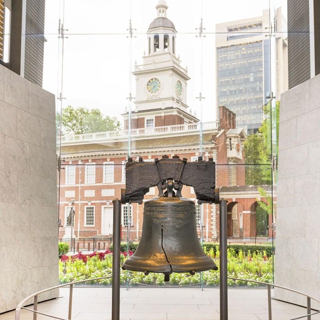 Color photo of the Liberty Bell with a red brick building visible through the glass wall behind it.
