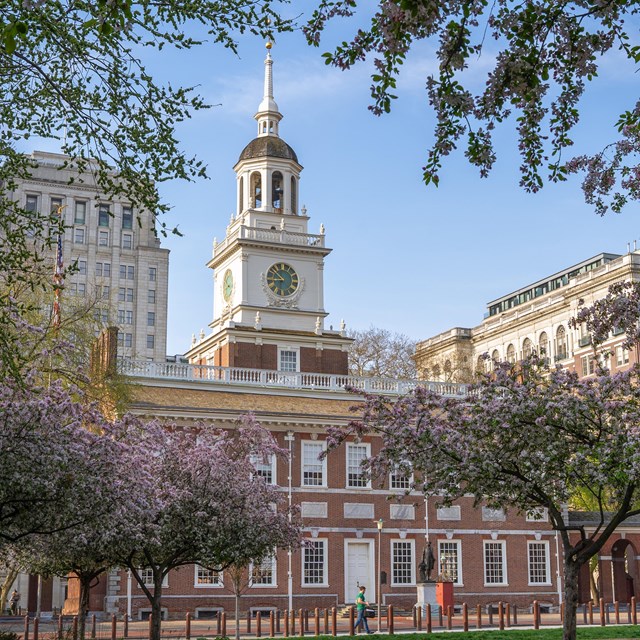 Two-story brick building with a white clock tower and cupola surrounded by trees in spring bloom