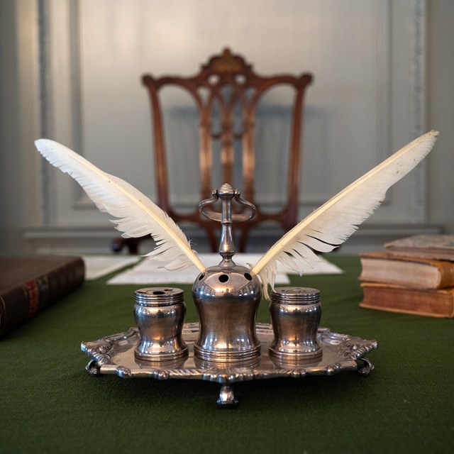 Silver inkstand with two white quill pens protruding on a table with a green table cloth.
