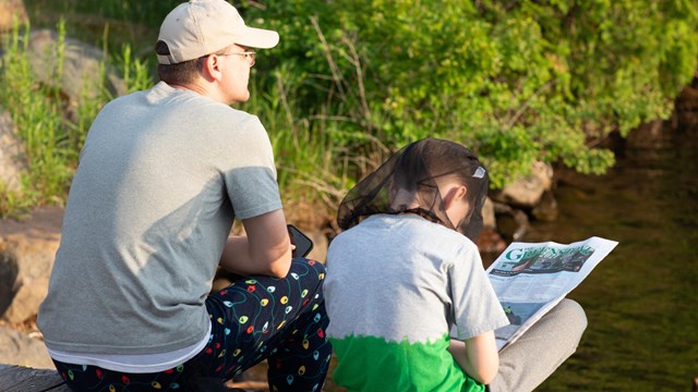 An adult and child sit on a dock looking out of the water while reading a newspaper.