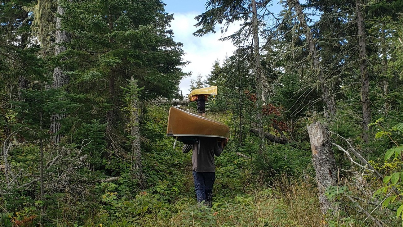 Two people, each portaging a canoe, hike on a trail up a hill surrounded by forest. 