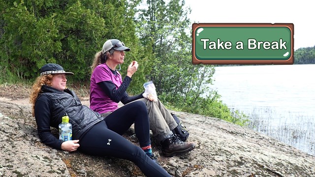 Two relaxed people lay on a rocky ridge near a lake.