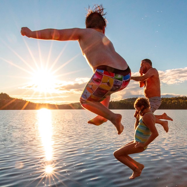 Three people jump from a dock into a lake. A fourth person watches from the dock.