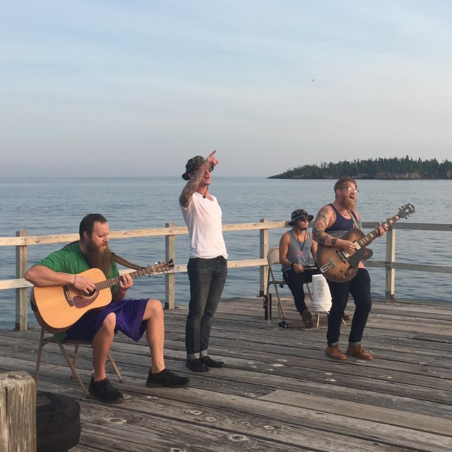 A photo shows four men playing music on a dock with water in the background