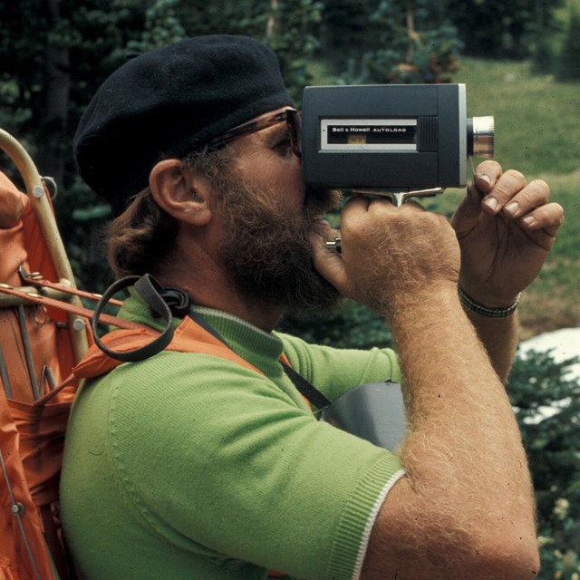 Image shows a man holding an old video camera with a large backpack on