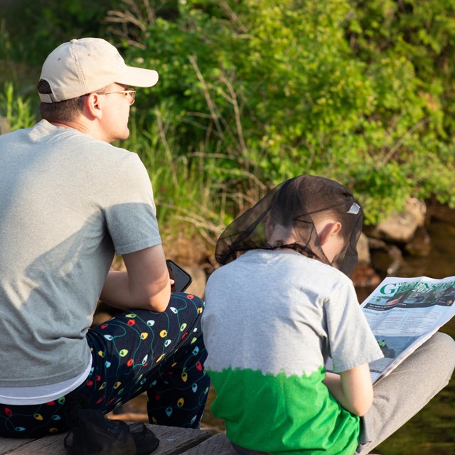 An adult and child sit on a dock looking out of the water while reading a newspaper.