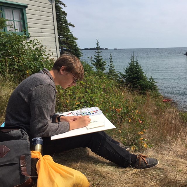 An artist works on a painting while sitting near a rocky lakeside.