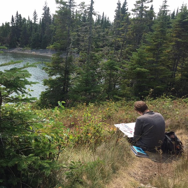 An artist works on a painting while sitting on the ground near a lake.