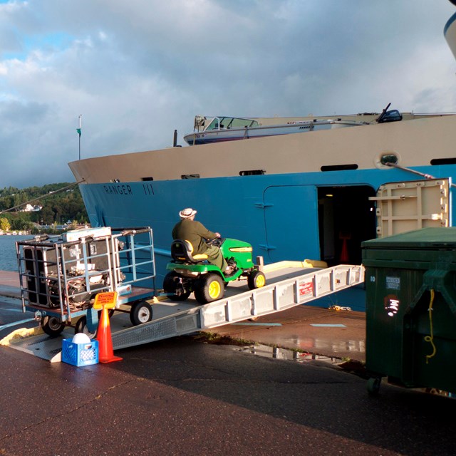 A tractor pulls two blue carts filled with baggage up a ramp into the cargo hold of RANGR III.
