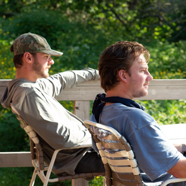 Two people sit in lawn chairs on a deck and look into the distance with green foliage behind them.