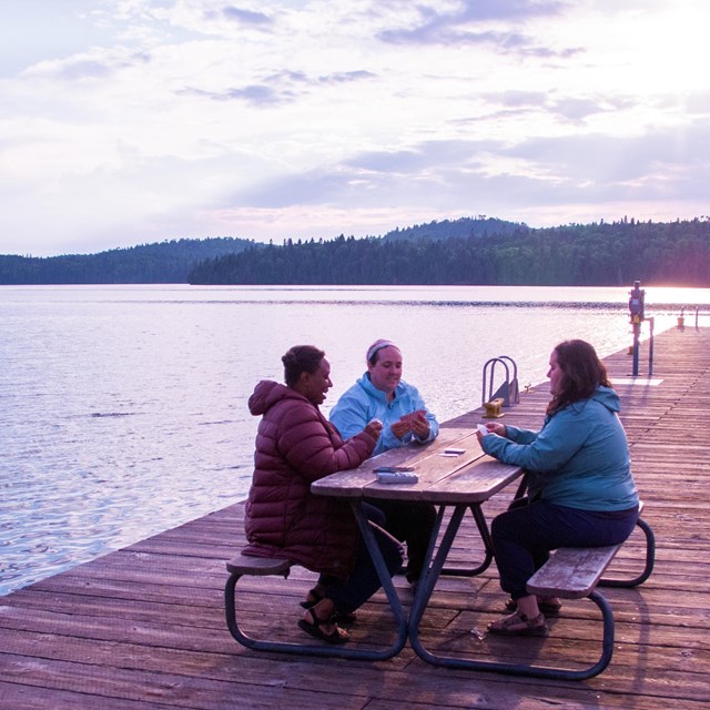 Three people sit at a picnic table on a dock playing cards while the sun sets behind them.
