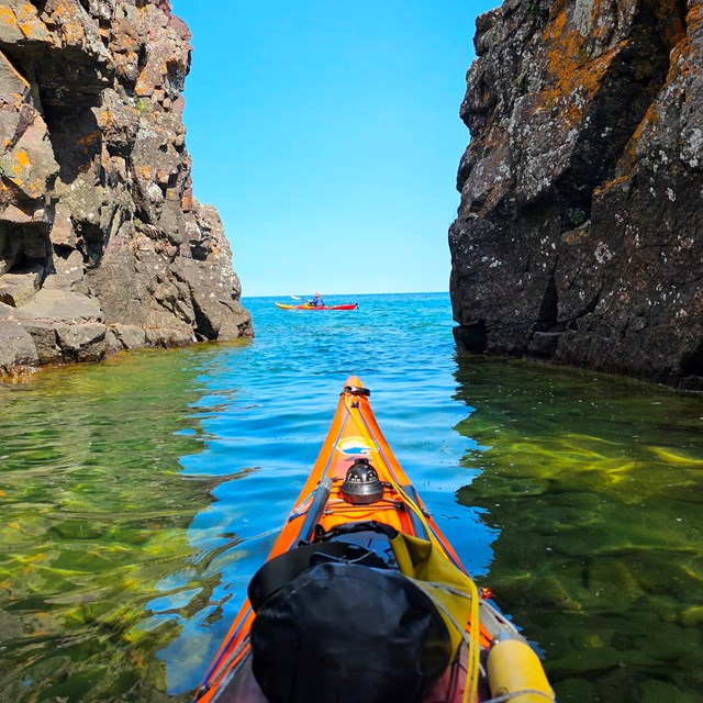 A yellow kayak maneuvers between two rocky cliffs on a calm sunny day in Lake Superior.
