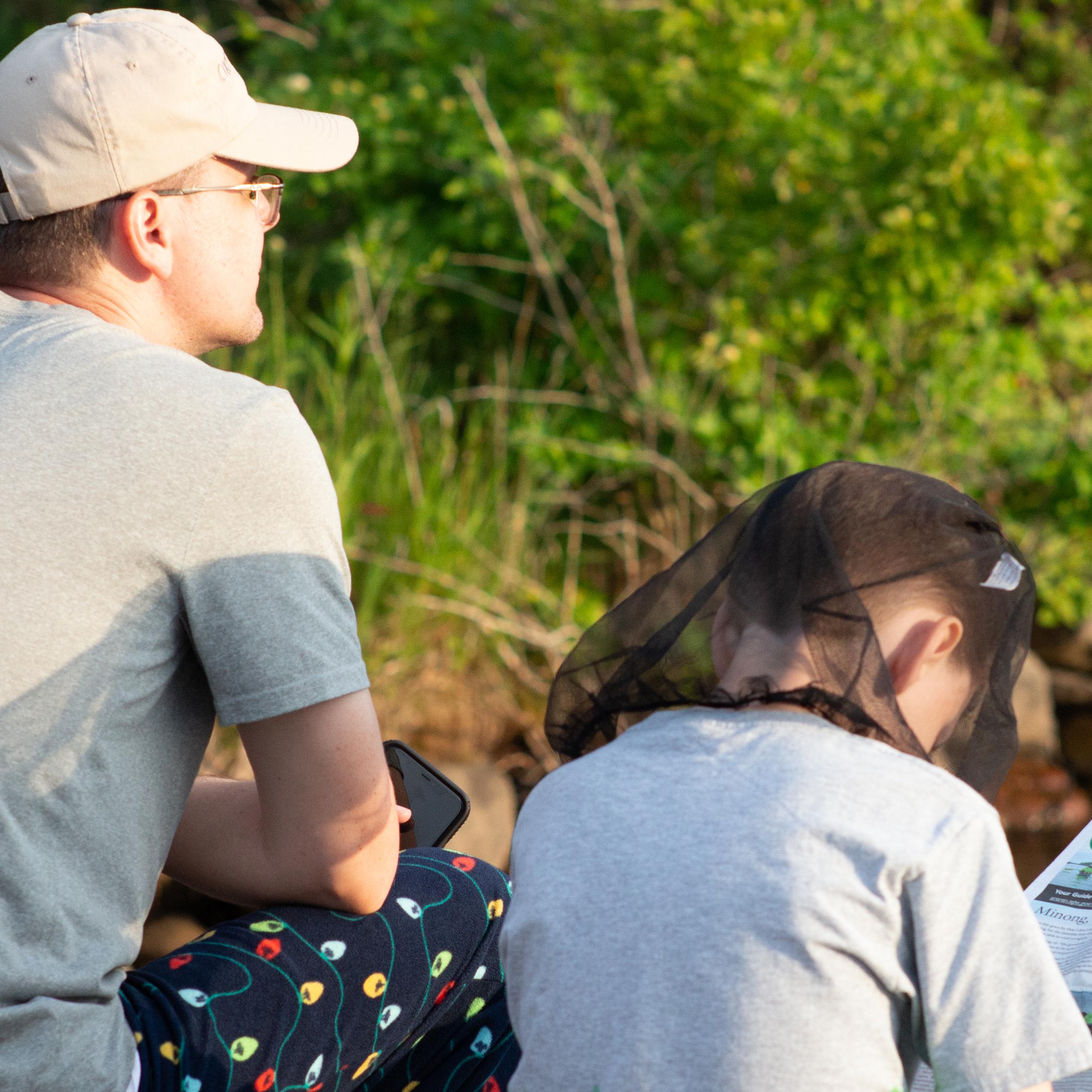 An adult sits on a dock with a child wearing a bug net.