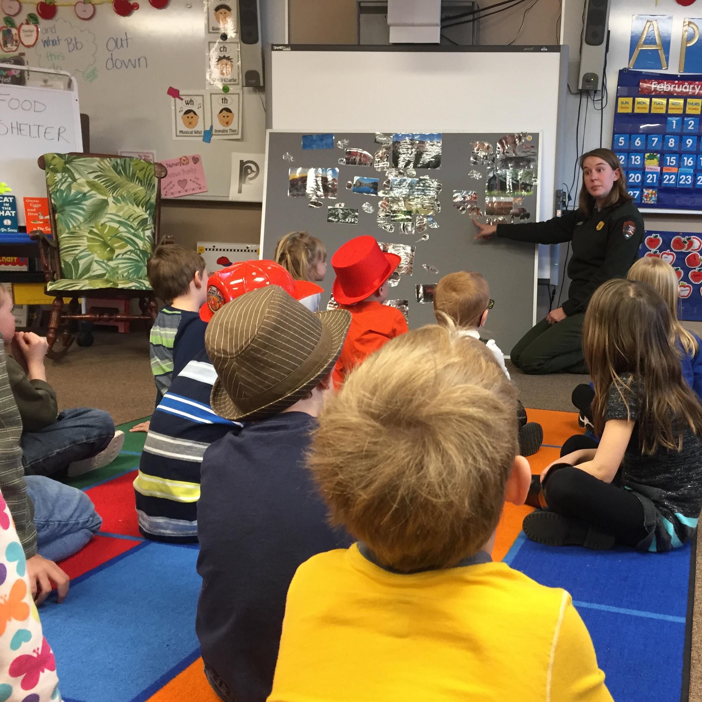 A ranger kneels in a classroom in front of several children while pointing at pictures of animals.