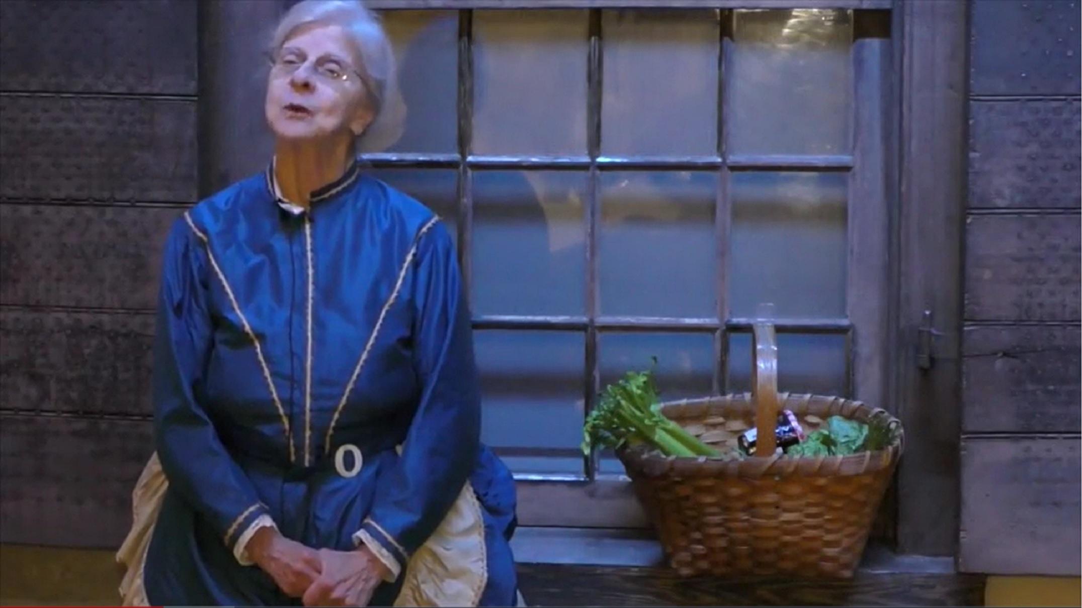 A woman in an old-fashioned blue dress stands next to a window with a basket of fresh vegetables