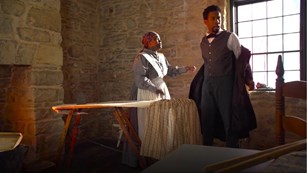 A man and woman in historic clothing stand next to an ironing board. The couple is African American.