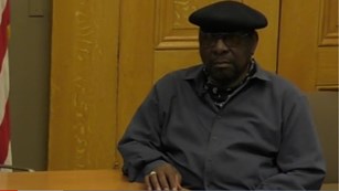 An African American man in a grey sweater and black cap sits next to a wooden table