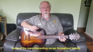 A park ranger sits on a leather couch holding a guitar. He has a white beard and mustache.