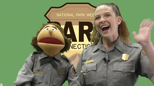 A ponytailed ranger with bright lipstick stands next to a puppet. Both are waving at the camera.