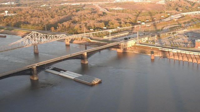 A tugboat pushing barges down a grey river. A shadow of an arch is reflected on the opposite bank.