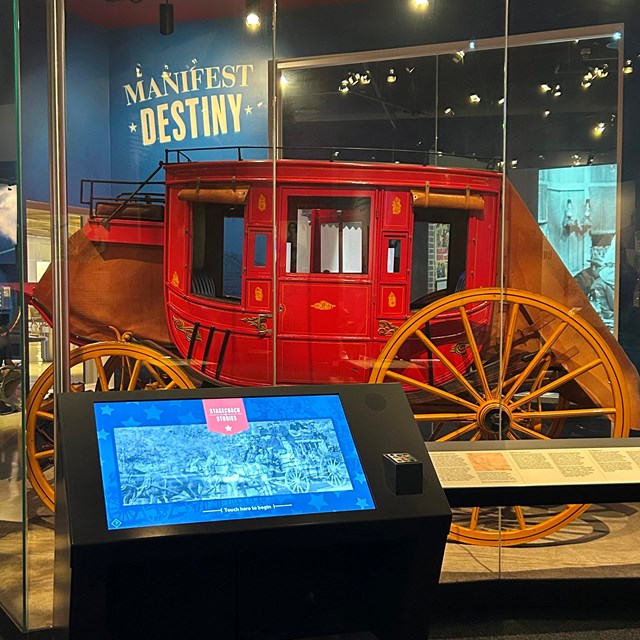 A red stagecoach with large yellow wheels behind glass at a museum
