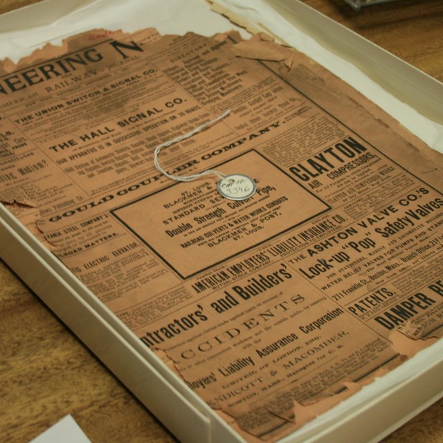 Yellowed, torn page from a newspaper or directory. Placed on a bed of paper inside an archival box.
