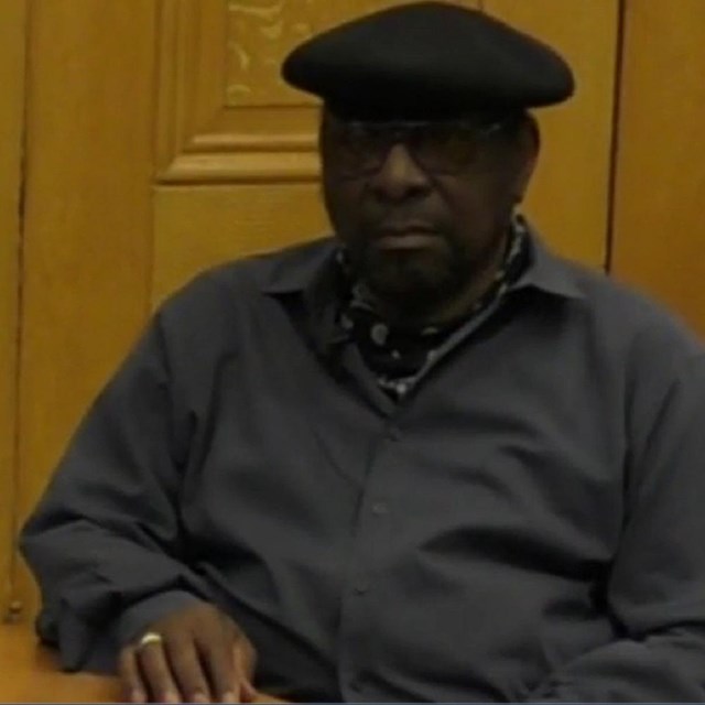 An African American man in a grey sweater and black cap sits next to a wooden table