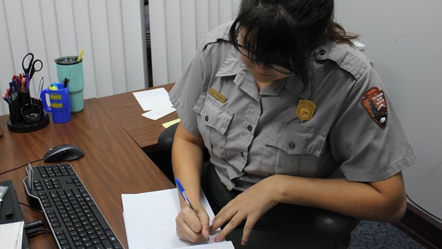 A woman in a gray button up with NPS patch sitting at a desk writing on paper.