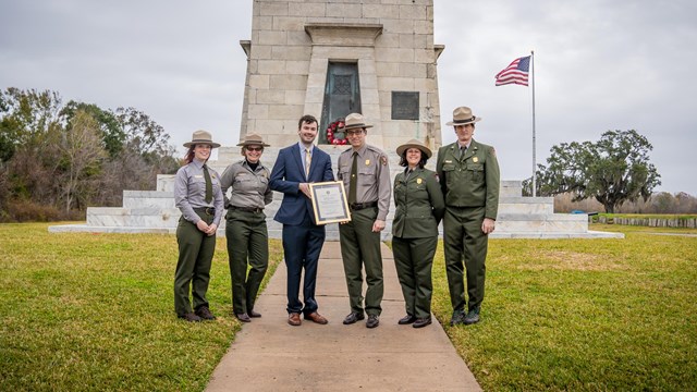 Park rangers stand in uniform in front of the Chalmette Monument, a tall white stone structure.