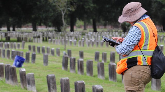 A woman in an orange safety vest looks at a clipboard and stands in a cemetery.