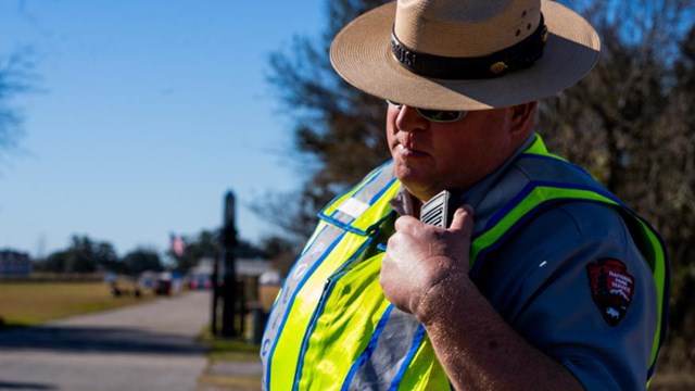A law enforcement ranger in a flat hat & wearing a safety vest talks into a radio