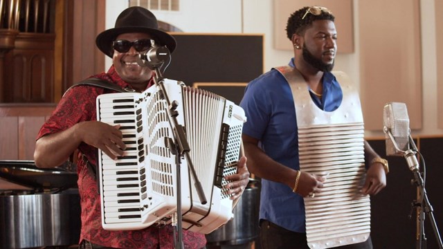 Two men are standing. One has a washboard on him. The other is holding an accordion.
