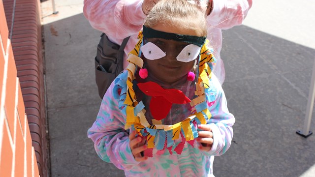 Outside on a sunny day a young girl smiles behind a mesh mask with felt face features on it.