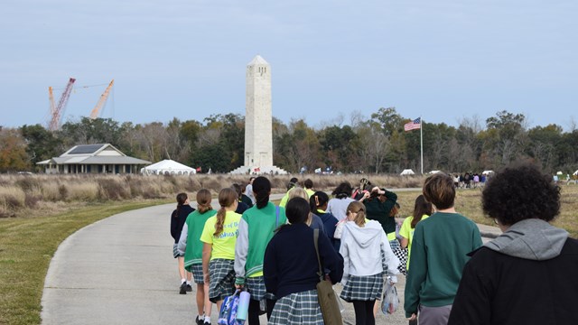 A group of students walk towards a tall stone monument and tents.