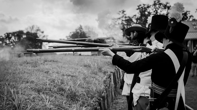 Black and white photo of men in living history clothes firing muskets.