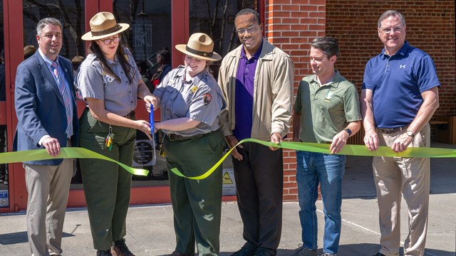 A line of people stand behind a green ribbon as two park rangers cut the ribbon with big scissors.