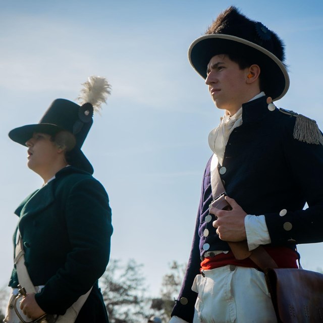 Two teenage boys stand outside in 1815 period clothing with hats, coats, and pantaloons.