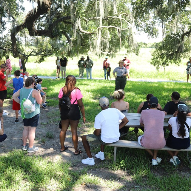 People standing around a Ranger giving a talk outside under some trees.