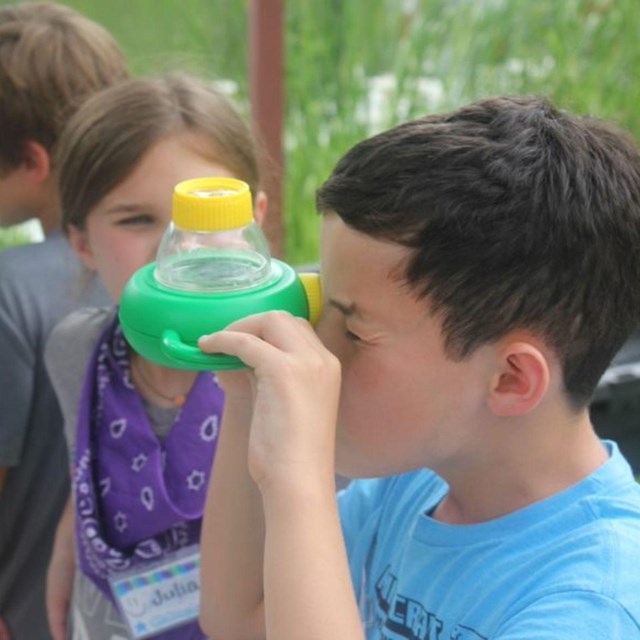 A young boy squints as he holds up a green magnifier to his eye.