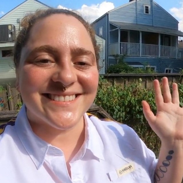 A girl in a white shirt is smiling and waving at the camera. There is a fence and house behind her.