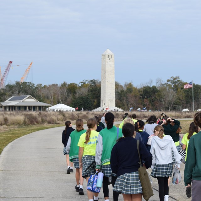 A group of students walk towards a tall stone monument and tents.