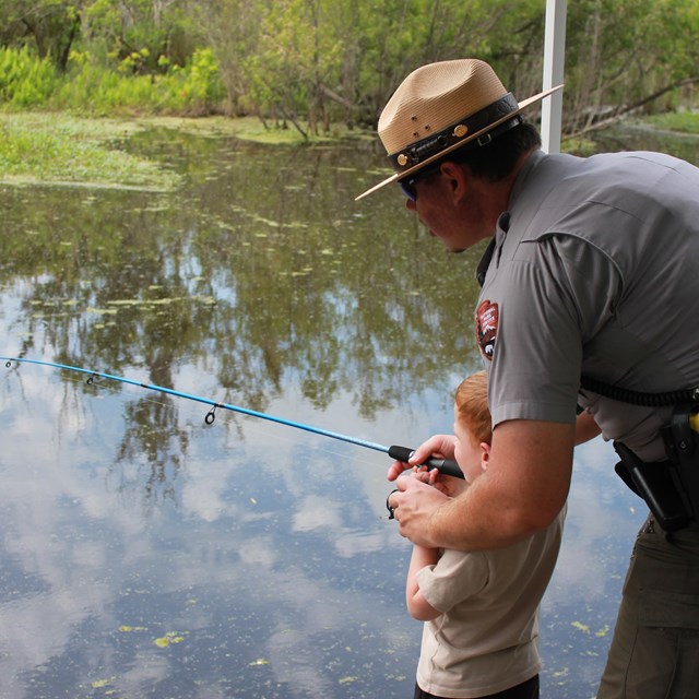 A law enforcement ranger helps a boy fish on the side of a bayou.