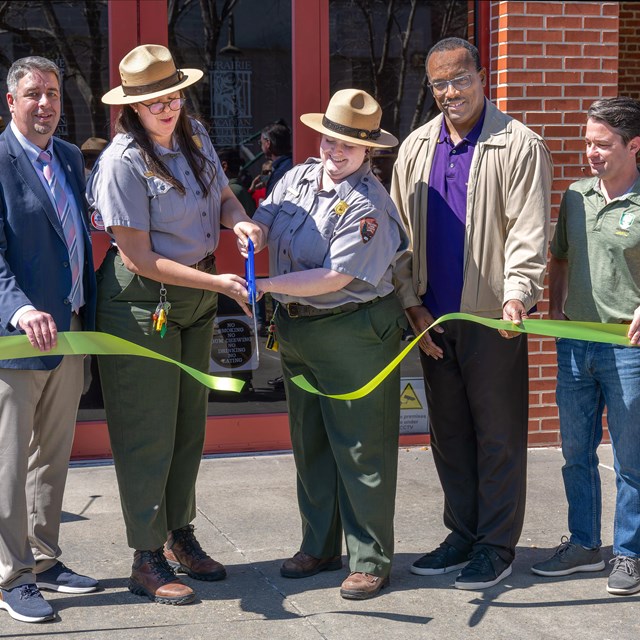 A line of people stand behind a green ribbon as two park rangers cut the ribbon with big scissors.
