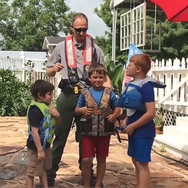 an adult with three kids in life jackets standing near a pool