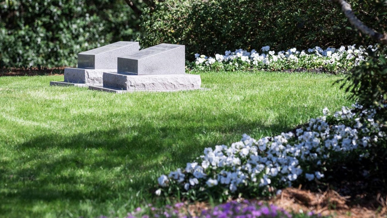 Two simple gray headstones side by side, surrounded by gardens of trees and flowers.