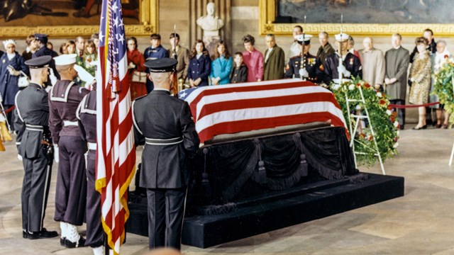 Casket covered by a flag with honor guard and wreaths