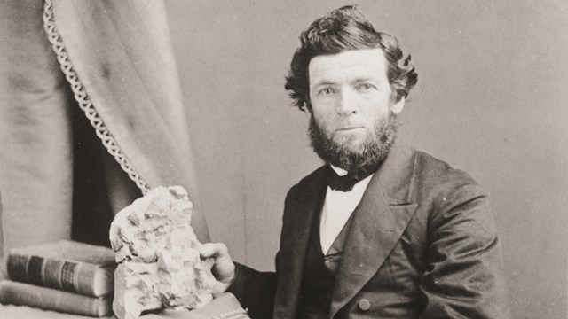 Black and white photo of a man with a beard in an old-fashioned suit and bow tie holding a rock.